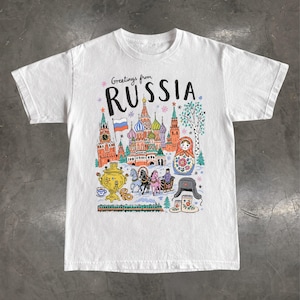 Może przedstawiać: Biały t-shirt z krótkim rękawem z kolorowym nadrukiem graficznym i napisem "Greetings from RUSSIA". Ilustracja przedstawia Sobór Wasyla Błogosławionego, samowar, matrioszkę, sanie ciągnięte przez konie, pociąg oraz zimowe elementy odzieży.