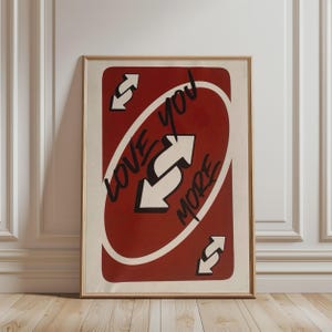 Op de afbeelding: Een rechthoekige wandkunstprint in een lichthouten lijst met een rood speelkaartontwerp met witte omkeerpijlen in de hoeken en in het midden. De zwarte tekst Love you MORE staat diagonaal over het midden van de kaart geschreven.