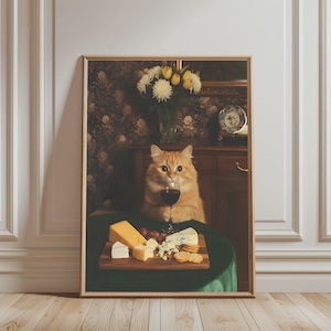 Könnte beinhalten: Ein gerahmter Wandkunstdruck mit einer flauschigen orangefarbenen Katze, die an einem runden Tisch mit dunkelgrünem Tuch sitzt. Die Katze hält ein Glas Rotwein. Auf dem Tisch steht ein Holzbrett mit verschiedenen Käsesorten, Crackern und Weintrauben.