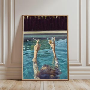 Könnte beinhalten: Ein gerahmter Wandkunstdruck, der eine Person zeigt, die in einem Swimmingpool untergetaucht ist und die Arme über das Wasser hebt. Eine Hand hält eine brennende Zigarette, die andere ein Martiniglas mit einer Olive. Im Hintergrund sind ein Pooldeck und Bäume zu sehen.