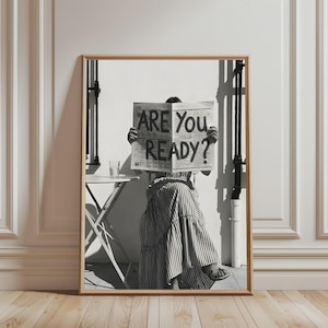May include: A black and white framed wall art print depicting a person sitting at a small table, holding a newspaper with the text ARE YOU READY? printed in large bold letters. The person wears a striped skirt and sandals in this outdoor scene.