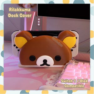 May include: A brown Rilakkuma dock cover for a gaming console, featuring a bear face with yellow inner ears, black eyes, and a white muzzle. The text Rilakkuma Dock Cover and Switch 1 / Oled Compatible is visible on the image, which shows the cover attached to a white console dock.