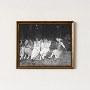 Pode incluir: Uma fotografia vintage a preto e branco emoldurada que retrata cinco pessoas vestidas com túnicas claras com asas de fada presas, sentadas ao ar livre numa zona arborizada. Cada pessoa segura um pequeno objeto e parece estar a fumar.