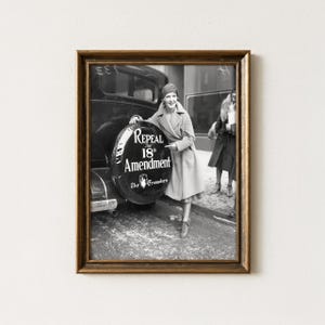 May include: A framed black and white photograph showing a person in a long coat standing next to a car with a spare tyre cover that reads Repeal The 18th Amendment The Crusaders. The image is enclosed in a simple brown wooden frame.