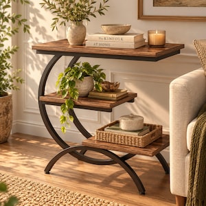 Peut inclure: Une table d'appoint à trois niveaux avec un cadre en métal foncé en forme de C et des étagères à grain de bois. L'étagère supérieure contient un vase, un bol, deux livres intitulés The Kinfolk At Home et une bougie. Une plante retombe.