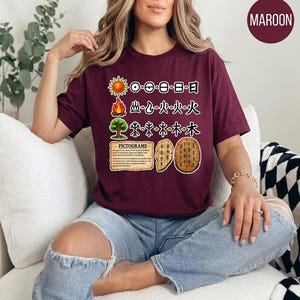 May include: A maroon t-shirt featuring a graphic print of three rows of pictograms showing the evolution of sun, fire, and tree symbols. Below the symbols is a text box titled PICTOGRAMS with two illustrations of ancient inscribed turtle shells.