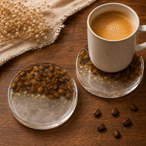 May include: Two circular resin coasters featuring embedded roasted coffee beans and gold leaf flakes. One coaster supports a ceramic mug filled with coffee, while the other sits empty. The coasters have a clear, glossy finish and a smooth surface.