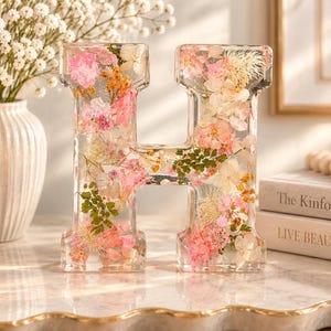 Può includere: Una lettera H decorativa in resina trasparente riempita con un assortimento di fiori secchi rosa, bianchi e gialli e fogliame verde. La lettera è autoportante e presenta una finitura liscia e lucida che mette in risalto gli elementi botanici.