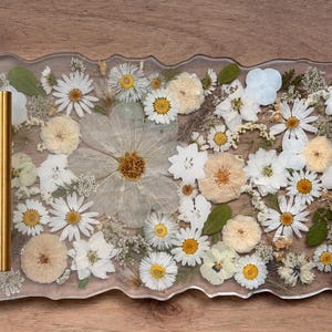 May include: A rectangular resin serving tray with an irregular, wavy edge, featuring a clear finish embedded with a variety of pressed white and cream-colored flowers and green leaves. The tray includes two vertical, brushed gold metal handles.