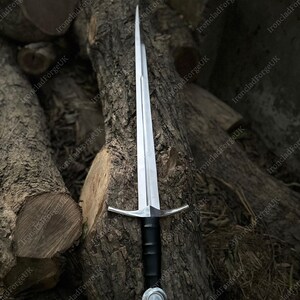 May include: A double-edged steel sword featuring a long, tapered blade with a central fuller, a straight crossguard, a black cylindrical grip, and a circular pommel. The sword is positioned vertically, showcasing its metallic finish and design.