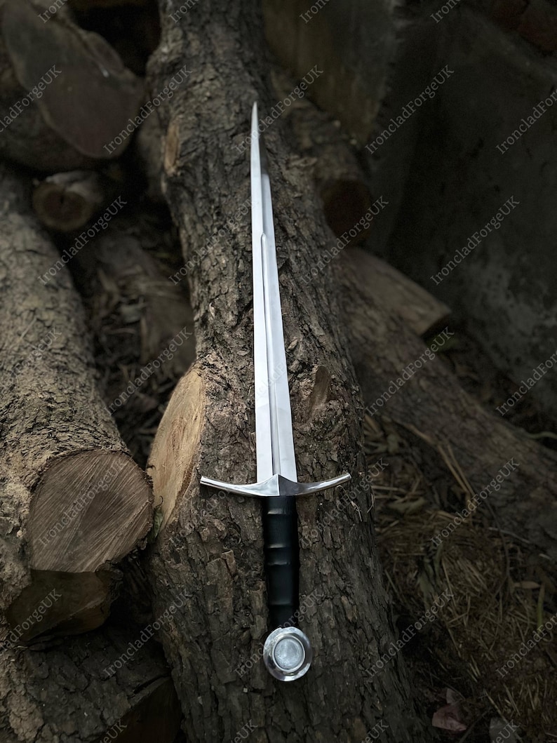 May include: A double-edged steel sword featuring a long, tapered blade with a central fuller, a straight crossguard, a black cylindrical grip, and a circular pommel. The sword is positioned vertically, showcasing its metallic finish and design.