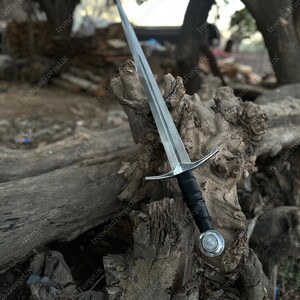 May include: A double-edged steel sword featuring a pointed blade with a central fuller, a straight silver-toned crossguard, a black wrapped grip, and a round silver-toned pommel with a central circular inlay, positioned upright.