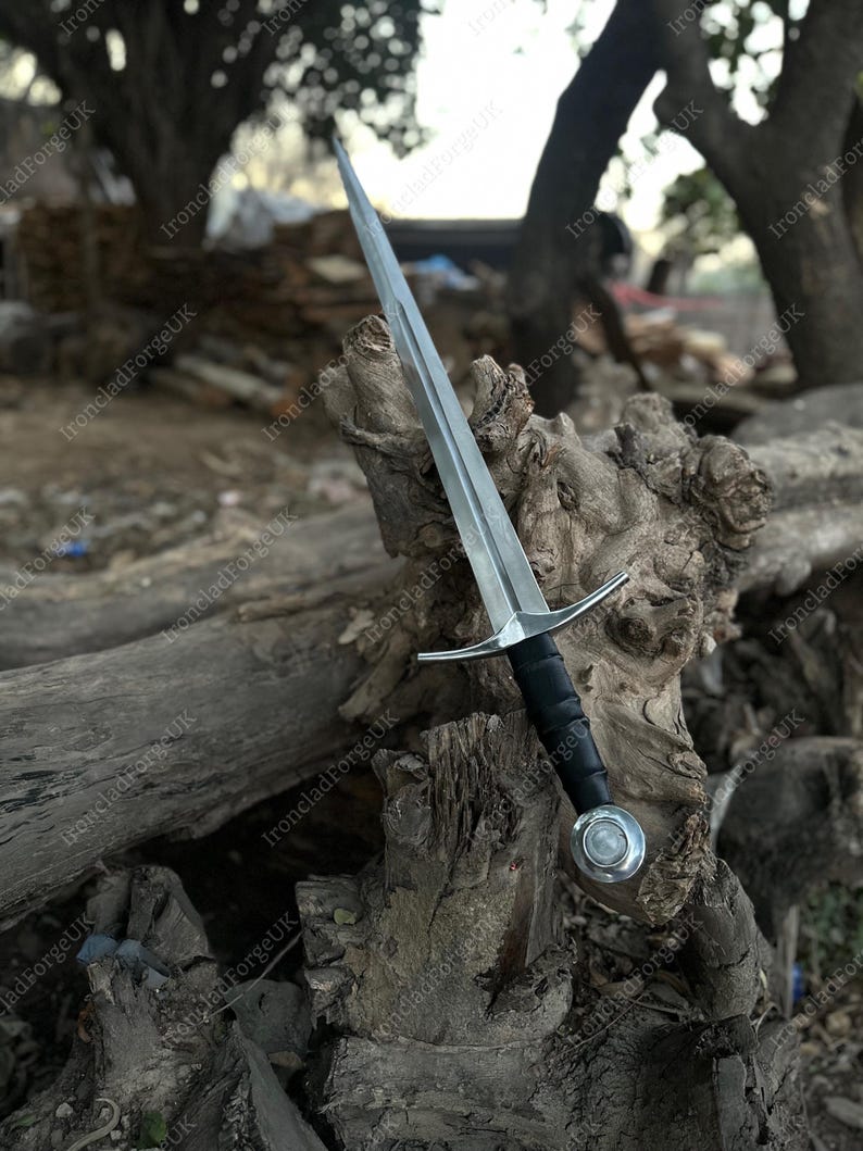 May include: A double-edged steel sword featuring a pointed blade with a central fuller, a straight silver-toned crossguard, a black wrapped grip, and a round silver-toned pommel with a central circular inlay, positioned upright.