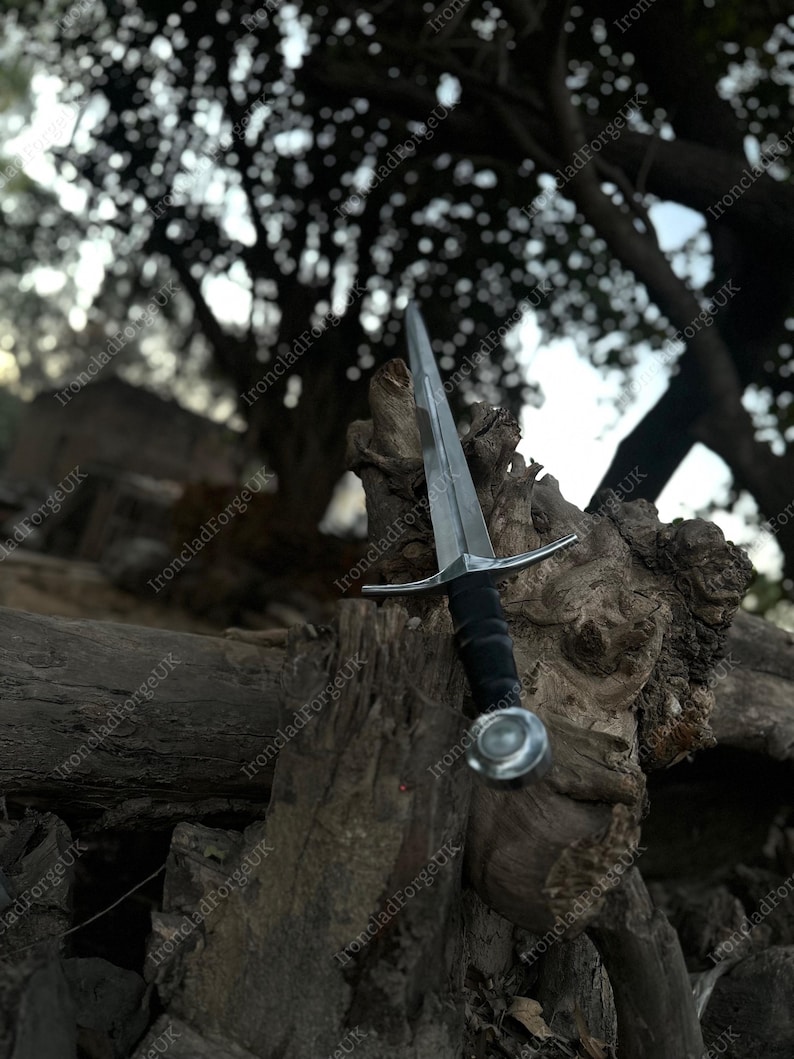 May include: A silver-colored metal sword with a straight, double-edged blade, a simple crossguard, and a black grip. The sword features a round, metallic pommel at the base of the handle, showcasing a clean, functional design for this weapon.