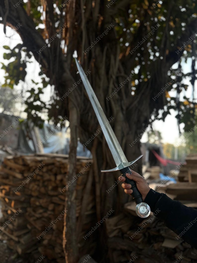 May include: A double-edged steel sword featuring a pointed blade, a straight crossguard, a black cylindrical grip, and a circular pommel with a central metallic disc. The sword is held upright, showcasing its metallic finish and construction.