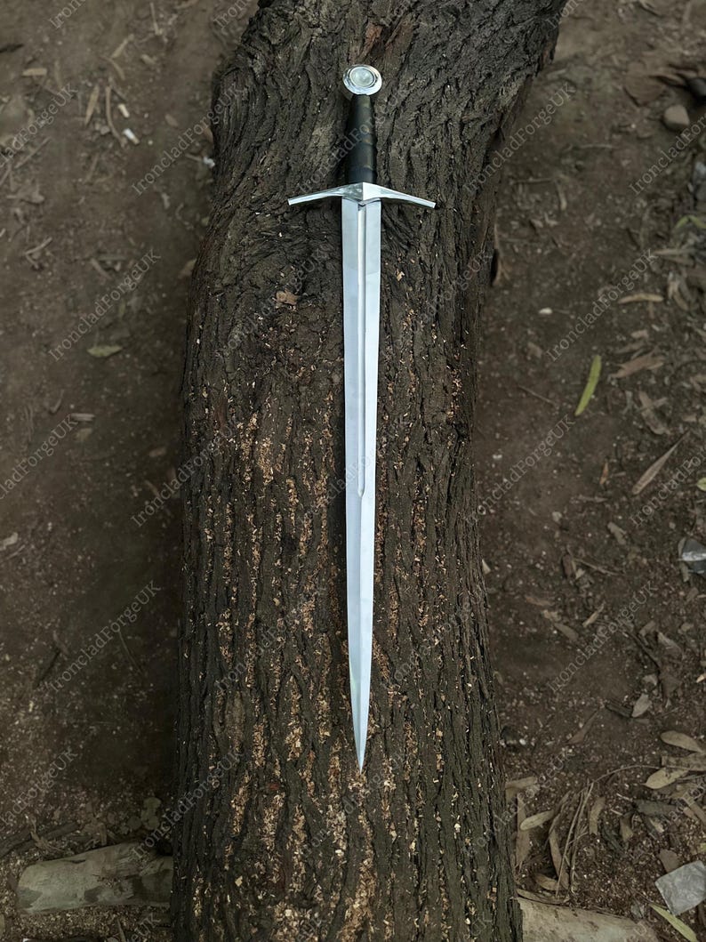 May include: A double-edged steel sword featuring a long, tapered blade with a central fuller, a straight silver-toned crossguard, a black leather-wrapped grip, and a round, polished silver-toned pommel at the end of the handle.