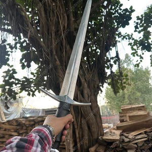 May include: A double-edged steel sword with a central fuller, a straight crossguard, a black grip, and a round pommel. The blade is pointed and metallic silver in color. The sword is held upright against a backdrop of trees and stacked wood.