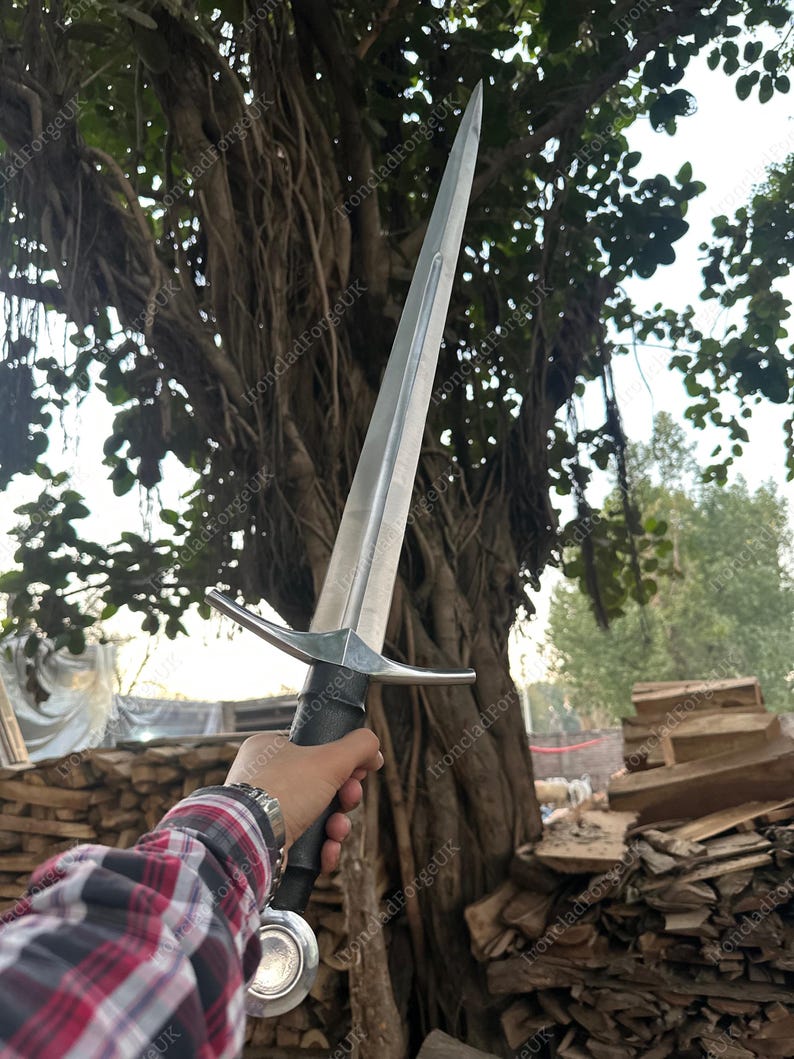May include: A double-edged steel sword with a central fuller, a straight crossguard, a black grip, and a round pommel. The blade is pointed and metallic silver in color. The sword is held upright against a backdrop of trees and stacked wood.