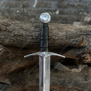 May include: A steel sword featuring a polished silver-colored blade, a straight crossguard, and a black leather-wrapped grip. The hilt is finished with a circular, flat-topped pommel. The design includes a central fuller running down the blade.