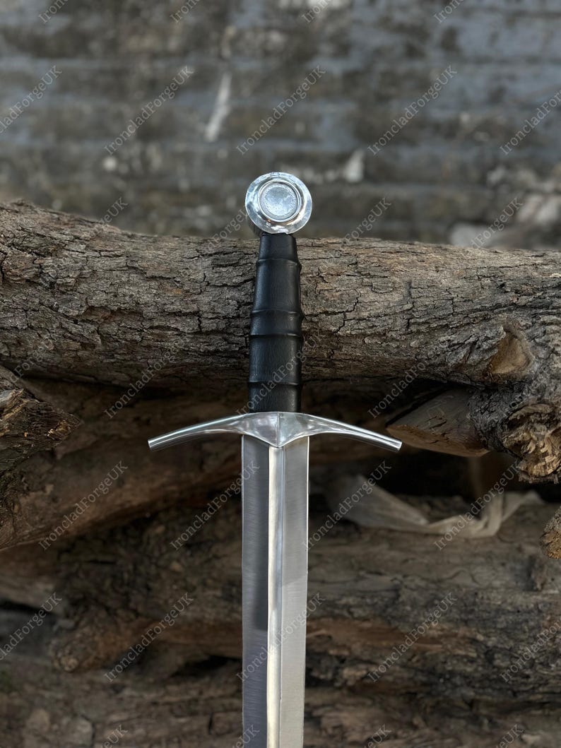 May include: A steel sword featuring a polished silver-colored blade, a straight crossguard, and a black leather-wrapped grip. The hilt is finished with a circular, flat-topped pommel. The design includes a central fuller running down the blade.