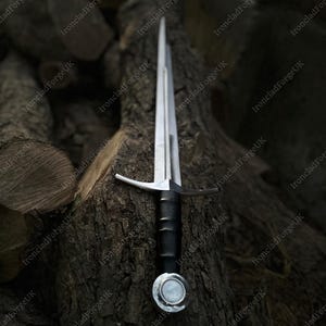May include: A double-edged steel sword featuring a long, pointed blade, a straight silver-toned crossguard, a black cylindrical grip, and a circular pommel with a central silver disc, positioned vertically along a textured wooden log.