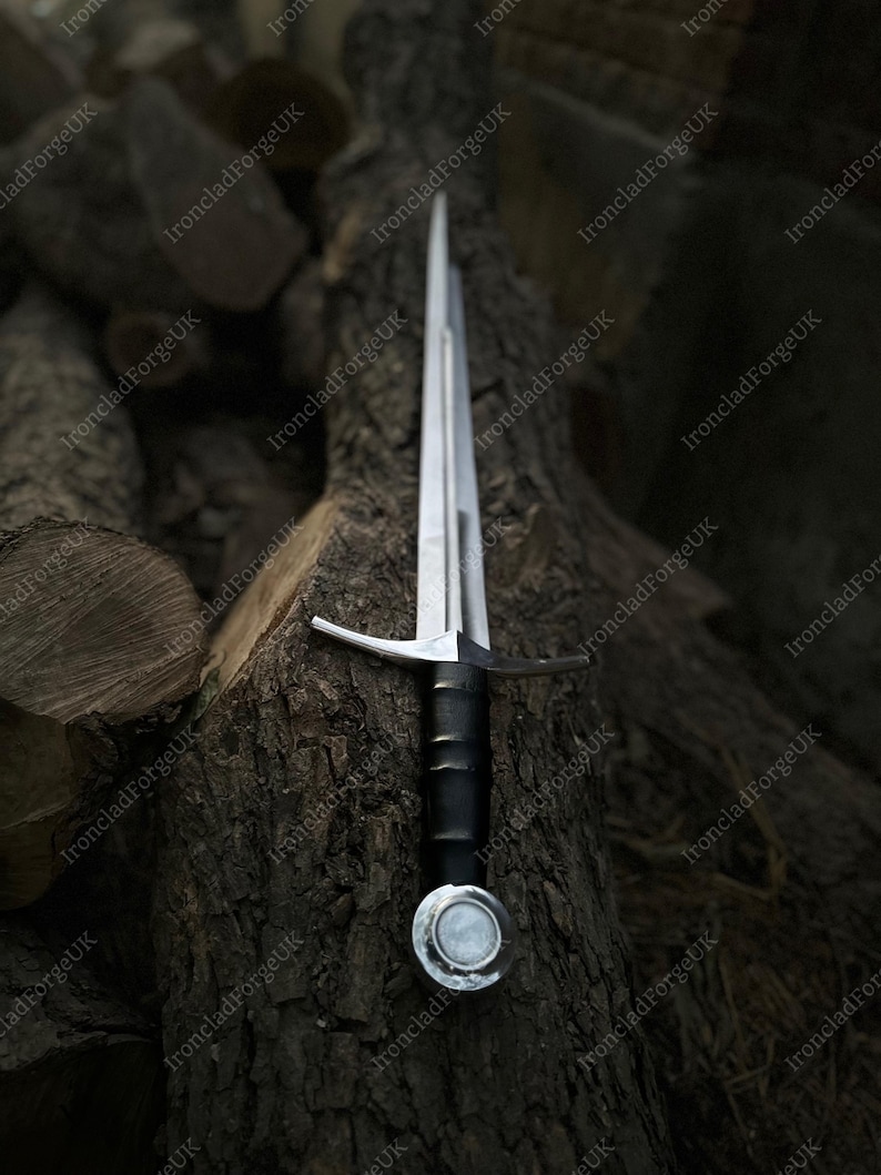 May include: A double-edged steel sword featuring a long, pointed blade, a straight silver-toned crossguard, a black cylindrical grip, and a circular pommel with a central silver disc, positioned vertically along a textured wooden log.