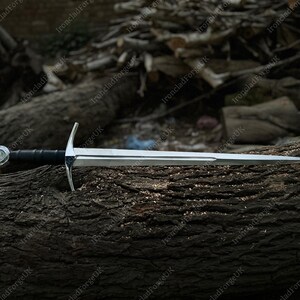 May include: A silver-colored metal sword with a double-edged blade, a straight crossguard, and a black leather-wrapped grip. The pommel is circular and metallic, featuring a central circular emblem. The sword is positioned horizontally.