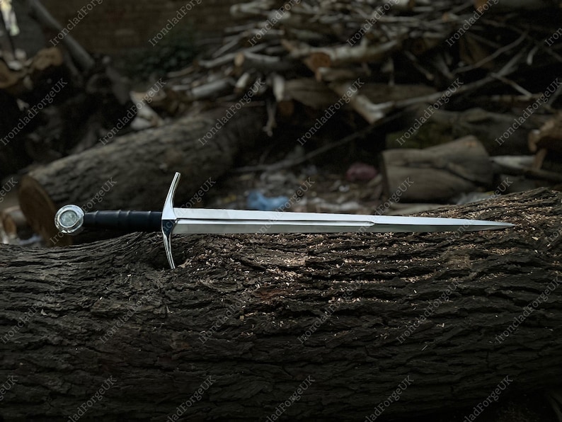 May include: A silver-colored metal sword with a double-edged blade, a straight crossguard, and a black leather-wrapped grip. The pommel is circular and metallic, featuring a central circular emblem. The sword is positioned horizontally.