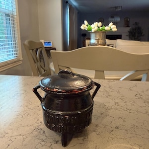 May include: A small, black, three-legged cast iron pot with a matching lid and two side handles. The exterior surface features embossed text that is partially worn, showing signs of age and use with some reddish-brown metallic oxidation.