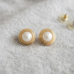 May include: A pair of round stud earrings featuring a central white faux pearl set within a polished gold-tone circular frame. The earrings have a smooth, metallic finish and include clear plastic earring backs for secure attachment to the ear.