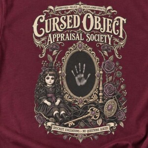 May include: A maroon t-shirt featuring a graphic print with the text "Cursed Object Appraisal Society," "Antiquities, Relics, Unexplained Effects," and "Discreet Evaluations, No Questions Asked." The design includes a doll, a mirror, and a key.
