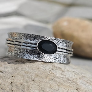 May include: A wide silver band ring featuring an oval-shaped black gemstone set in the centre. The metal surface is decorated with an intricate, embossed floral pattern and horizontal grooved lines that run across the entire circumference.