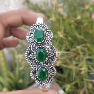 Può includere: Un bracciale color argento con tre pietre preziose verdi ovali e sfaccettate allineate verticalmente. La montatura in metallo presenta complessi motivi a volute, dettagli a perline e una trama a corda intrecciata attorno a ogni pietra.