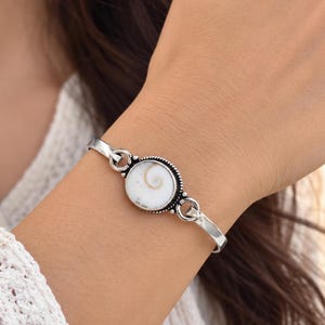 May include: A silver-toned metal cuff bracelet featuring a central circular setting. The centrepiece contains a white, flat, round shell with a natural brown spiral pattern. The metal frame around the shell has a beaded, textured edge detail.