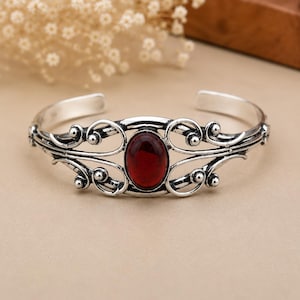 May include: A silver cuff bracelet featuring an oval-shaped, deep red gemstone at the centre. The metal band is decorated with intricate, swirling filigree patterns and small spherical accents, creating a symmetrical design across the front.
