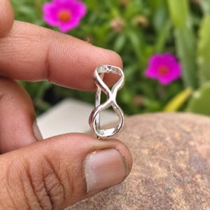 Könnte beinhalten: Ein Ring aus poliertem Silber mit einem Unendlichkeitssymbol-Design. Die Ringschiene besteht aus einem durchgehenden Metallstück, das zu einer Acht geformt ist und eine symmetrische, offene Struktur bildet, die zwischen zwei Fingern gehalten wird.
