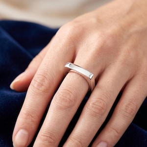 Könnte beinhalten: Ein polierter Ring in Silberoptik mit einer flachen, rechteckigen Oberfläche. Die Ringschiene ist glatt und metallisch, mit einem sauberen, reflektierenden Finish, das das Licht einfängt. Das Design ist schlicht und besteht aus einem Balken.