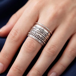 May include: A stack of five silver rings worn on a finger. The set includes a plain polished band, a band with an intricate etched pattern, a band with a beaded texture, and a band featuring diagonal line engravings, all crafted in a metallic silver tone.