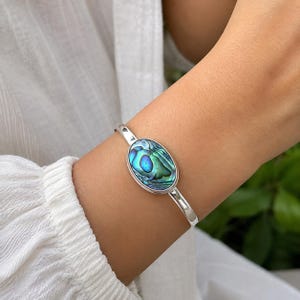 Pode incluir: Uma pulseira de prata tipo bracelete com uma incrustação central oval de concha de abalone. A concha exibe redemoinhos iridescentes em tons de azul, verde e roxo. A banda de metal é polida e lisa, envolvendo o pulso com um design simples.