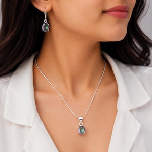 May include: A silver necklace and matching drop earrings set featuring oval-shaped iridescent stones. The necklace has a thin silver chain with a single oval pendant, and the earrings consist of matching oval stones hanging from silver hooks.