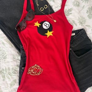 Pode incluir: Uma regata vermelha canelada com um gráfico de bola oito preta ladeado por duas estrelas amarelas. O conjunto inclui calças de ganga pretas, um colar dourado com pequenos brincos de argola, óculos de sol escuros e uma pulseira dourada.