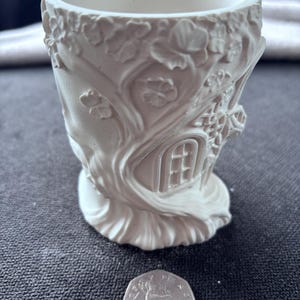 May include: A white cylindrical planter featuring a relief design of a tree with branches, leaves, and a small arched window. A British fifty pence coin is placed in front of the planter to provide a sense of scale for the object's size.