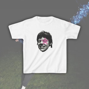 May include: A white short-sleeved t-shirt featuring a black and white photographic print of a person's face centered on the chest. A pink star graphic is positioned over the right eye of the face in the print, adding a distinct color accent.
