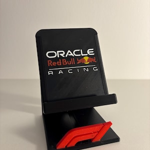 Peut inclure: Un support pour smartphone noir arborant le logo Oracle Red Bull Racing en blanc, rouge et jaune. Le support comprend un petit emblème rouge avec le logo F1 fixé sur la base, offrant une structure de soutien stable pour un appareil mobile.