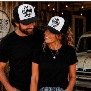 May include: Two black and white trucker hats featuring mesh backs and adjustable snap closures. The left hat displays the text I'M DUMB STUFF, while the right hat displays the text I DO DUMB STUFF. Both hats have a white front panel and black brim.