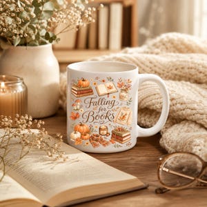 May include: A white ceramic mug featuring a colorful autumn-themed illustration of pumpkins, stacks of books, an open book, a sweater, a scarf, and a candle, with the text "Falling for Books" printed in a brown script font on the front surface.