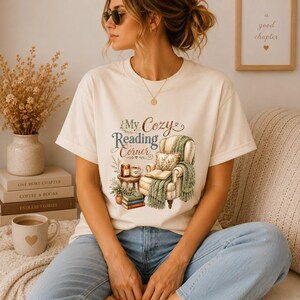 May include: A cream-colored short-sleeve t-shirt featuring a graphic illustration of an armchair with a green throw blanket, a stack of books, and a candle. The text "My Cozy Reading Corner" is printed above the armchair in brown and blue script.