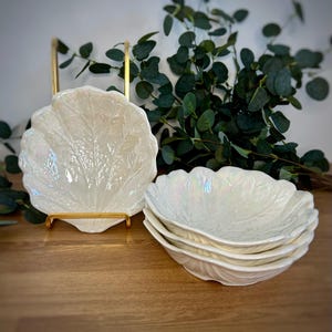 May include: A set of four white ceramic bowls with an iridescent finish and a textured leaf-vein pattern. One bowl is displayed upright on a gold metal stand, while the other three are stacked together in a nested pile on a wooden surface.