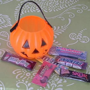 May include: An orange plastic pumpkin-shaped trick-or-treat bucket filled with various candy bars, including Milky Way, Snickers, Musketeers, Reese's Peanut Butter Cups, and Skor.