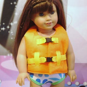 May include: A doll wearing an orange life vest with yellow straps and blue and white patterned swim trunks. The doll has long brown hair and blue eyes.